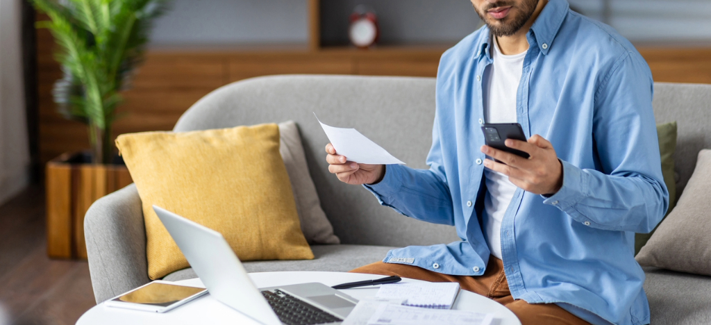 Leveraging electronic billing - A man at home reviewing his medical bill using his smartphone and laptop.