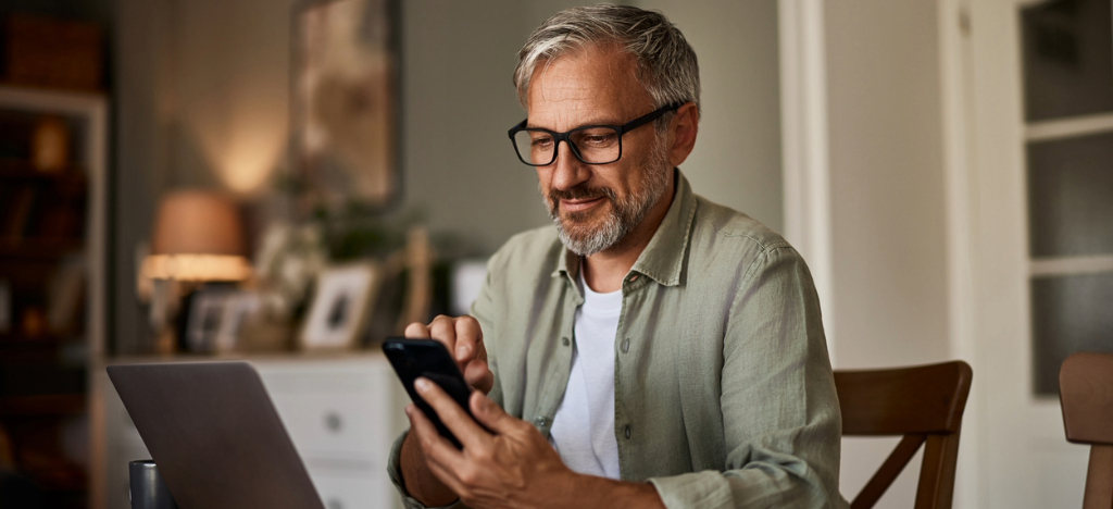 Improving payment reminder process - A senior man at home checking his hospital bill using his smartphone and laptop.