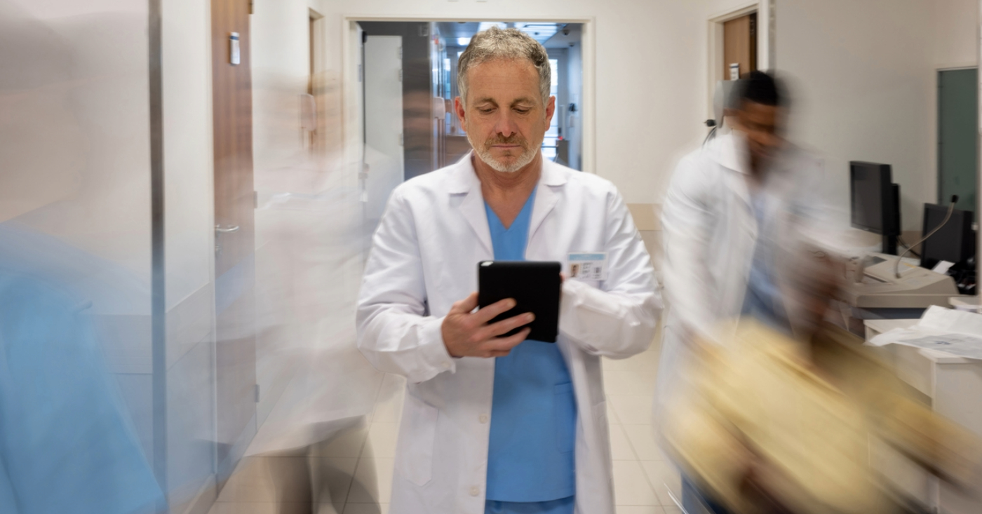 Managing patient surge - A male doctor holding a tablet device while standing in a busy hospital hallway.