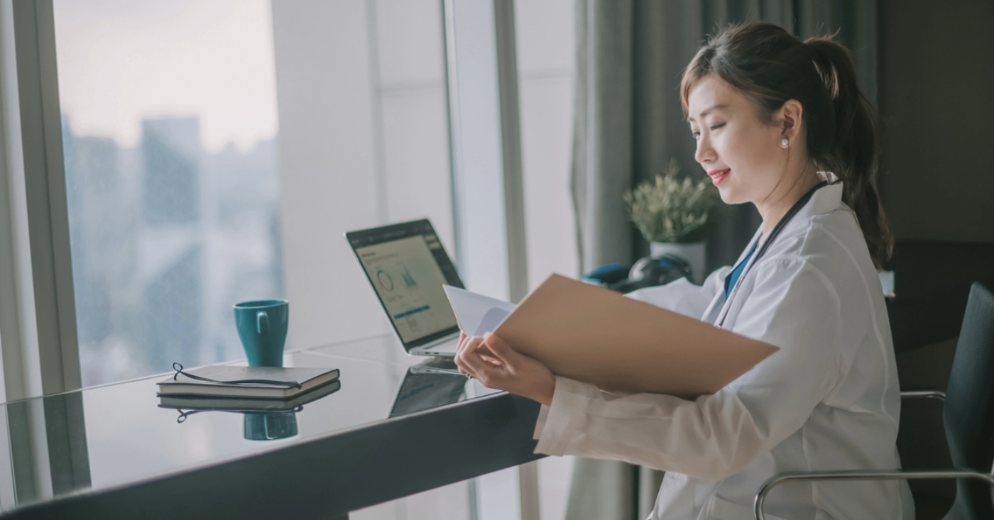 Electronic billing benefits - A female doctor looking at printed patient bills and using a laptop to access their practice’s medical billing software.