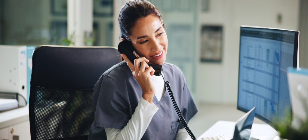 A female medical office staff member checking accounts using a computer while on the phone with a patient.