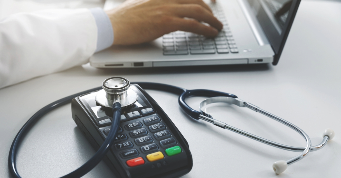 Stop chasing payments - A card reader and stethoscope on a table with a doctor working in the background.