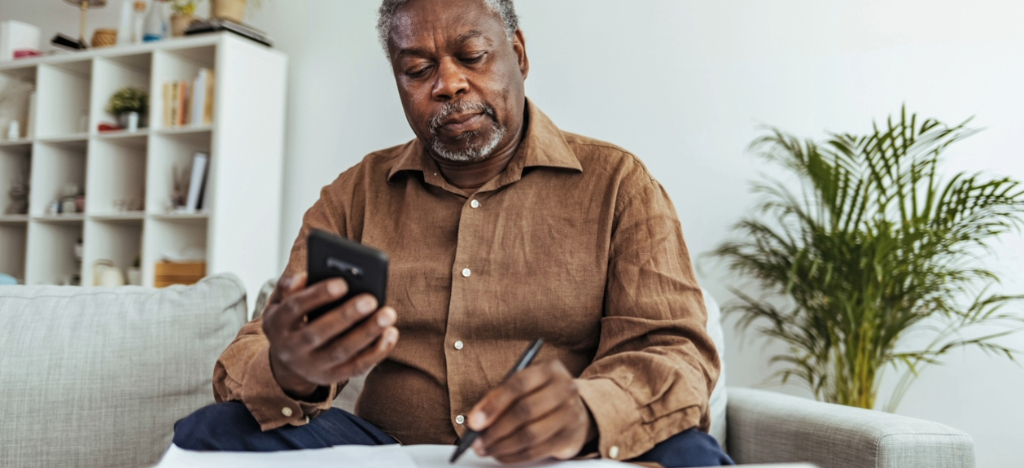 Improving billing experience - A senior man at home writing down his expected medical bill shown on his smartphone.